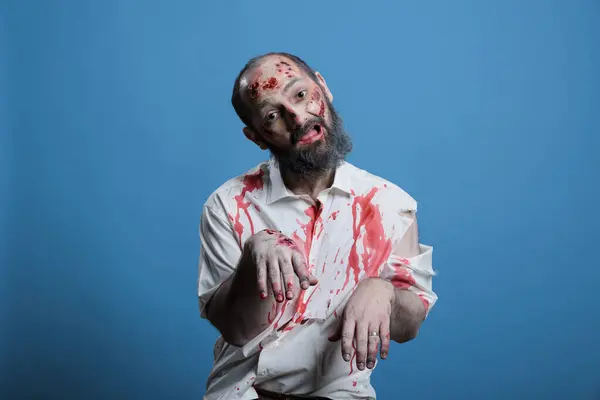 stock image Portrait of brainless zombie isolated over blue studio background limping around, looking clueless. Mindless reanimated corpse covered in blood and wounds looking at camera