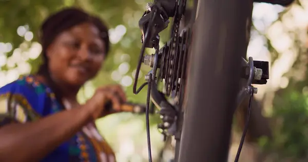 stock image Dedicated sporty african american woman checking performance of bicycle parts by spinning the pedals. Active female cyclist ensuring proper function of tire close-up of bike wheel in home yard.