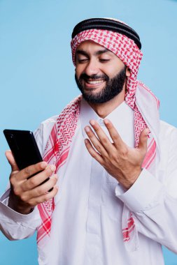 Gülümseyen Müslüman adam video konferansında akıllı telefon kullanıyor ve el hareketleriyle konuşuyor. Geleneksel giysiler içinde neşeli bir Arap cep telefonuyla görüntülü konuşma yapıyor.