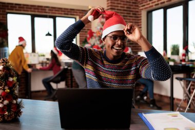 Neşeli bir çalışan Noel arifesinde Noel Baba şapkasını takıp Noel Baba 'nın masasında süslenmiş bir ofiste çalışırken kameraya bakıyor. Kış tatilinde işyerinde Afrikalı Amerikalı bir adam vardı.