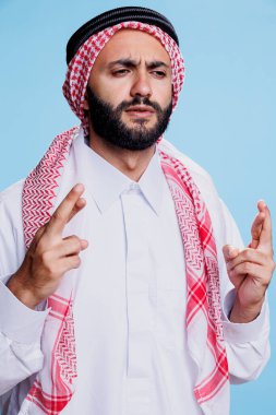 Kültürel tobe giymiş ve kaşlarını çatmış kaşlarını çatmış ve dilek tutarken parmaklarını çaprazlamış Arap bir adam. Batıl inançlı müslüman, şans diliyor.