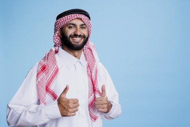 Geleneksel giyinmiş Arap adam stüdyoda başparmağıyla poz verirken gülümsüyor, müslüman kültür portresi gösteriyor. Gülümseyen neşeli İslami kişi onay jesti yapıyor ve kameraya bakıyor.