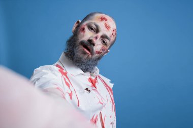 Gerilim filmi zombisi gibi giyinmiş aktör portresi sette sahneler arasında selfie çekiyor. Adam kanlı makyajla kaplı yürüyen ölü bir yaratık gibi giyinmiş. Fotoğraf çekiyor, stüdyo arka planında izole edilmiş.