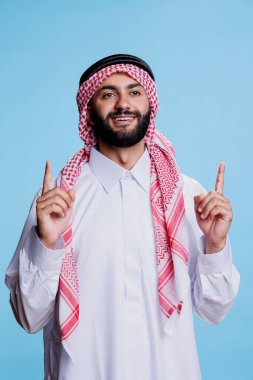 Geleneksel tobe ve ghutra gibi giyinmiş, iki parmağıyla portreyi gösteren neşeli Arap adam. Ürün reklamları yaparken Arap cübbesi giyen müslüman bir kişi ortaya çıkıyor