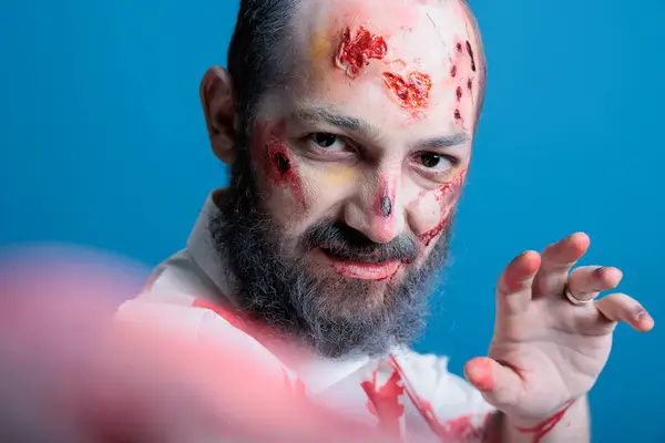 stock image Man dressed as zombie for Halloween event turning hand into claw, throwing ferocious look. Person costumed as undead creature covered in blood makeup making himself look dangerous, studio background