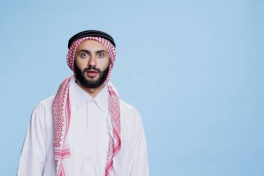 Geleneksel giyinmiş Müslüman adamın kameraya geniş gözlerle bakmasına şaşırdım. Arap kişi, şok olmuş yüz ifadesi portresiyle poz verirken başörtüsü ve başörtüsü takıyor.