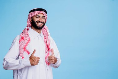 Geleneksel cübbe ve başlık giymiş gülümseyen müslüman adam başparmağıyla hareket portresi yapıyor. Neşeli Arapça onay işareti gösteriyor ve kaygısız bir ifadeyle kameraya bakıyor.
