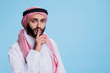 Müslüman adam geleneksel kıyafetler giyiyor sessiz bir jest yapıyor ve dudak portresi üzerinde işaret parmağı tutuyor. İslami başörtüsü takmış bir Arap sessiz bir işaret veriyor ve kameraya bakıyor.
