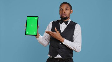 Bellman hotelier showing a tablet with green screen display in studio, presenting mockup chroma key screen against blue background. Young hotel concierge works at resort. Camera B. clipart