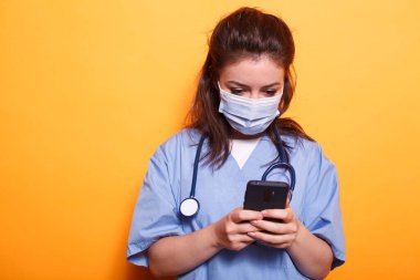 Caucasian female doctor utilizing modern technology while wearing face mask against coronavirus. Medical assistant with stethoscope texting on her mobile phone during pandemic. clipart