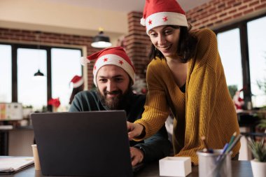 Noel Baba şapkalı gülümseyen adam ve kadın iş arkadaşları dizüstü bilgisayarda çalışıyor ve ofiste Noel 'i kutluyorlar. Şenlikli işyerindeki bilgisayar projesini yöneten neşeli meslektaşlar