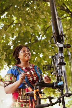 Excited sports-loving african american woman conducting annual summer bike maintenance. Energetic young female cyclist inspecting and ensuring proper functioning of modern bicycle. clipart