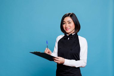 Gülümseyen Asyalı garson üniforma giyiyor, pano tutuyor, kalemle yazıyor ve kameraya bakıyor. Neşeli kadın resepsiyonist mavi arka planda stüdyo resmi çekiyor.