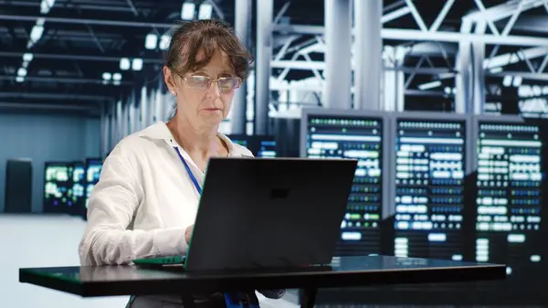 stock image Qualified worker in server farm housing high end rows capable of quickly and efficiently performing complex computations and data analysis, using mockup laptop to get rid of problems