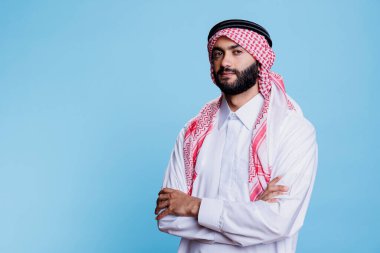 Geleneksel giysiler giymiş, kolları çaprazlanmış, kendinden emin Müslüman bir adam. Tobe ve ghutra giymiş genç Arap adam katlanmış elleriyle poz veriyor ve kameraya bakıyor.