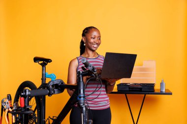 Enthusiastic sports-loving black woman with laptop repairing bicycle in front of yellow background. Healthy african american lady searching on personal computer to fix broken bike components. clipart