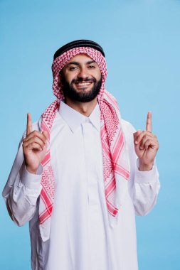 Arap adam geleneksel İslami cübbe ve başörtüsü giyiyor. Üzerinde parmak stüdyo portresi var. Thombe ve ghutra giymiş gülümseyen müslüman yukarı bakıyor ve kameraya bakıyor.