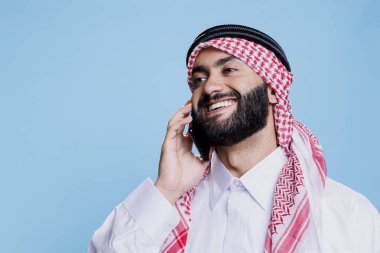 Geleneksel kıyafetli neşeli Müslüman adam akıllı telefonla konuşurken gülüyor ve stüdyoda poz veriyor. Damalı ghutra ile gülümseyen Arap başörtüsü telefonuna mutlu bir ifadeyle cevap veriyor.