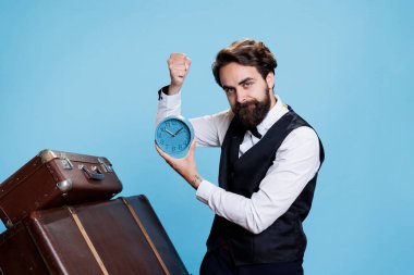 Hotel employee looks at time on camera, trying to be punctual in hospitality industry with wall clock. Professional doorkeeper in suit and tie appearing with watch against blue background. clipart