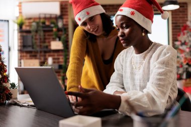 Afrika kökenli Amerikalı kadın ofis çalışanı Noel arifesinde dizüstü bilgisayarla yöneticiye rapor gösteriyor. Noel Baba şapkası takan ve kış tatilini iş yerinde kutlayan şirket çalışanları.