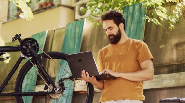 Kırık modern bisikleti çevrimiçi elle tamir etmek için dizüstü bilgisayarını kullanan sağlıklı, beyaz bir adam. Spor düşkünü erkek bisikletçi elinde mini bilgisayarla dışarıda bisiklet bakımı için talimat alıyor..