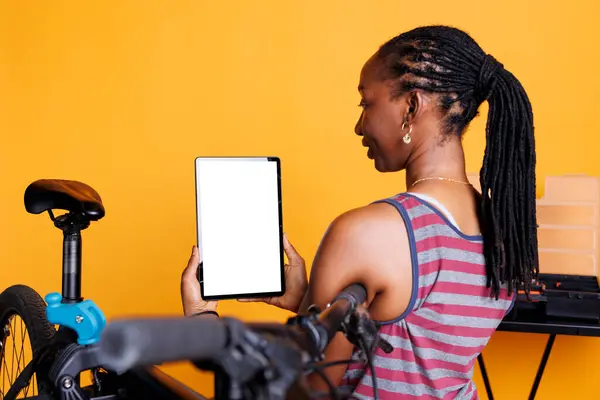 stock image Detailed shot of black woman carrying a device displaying blank copyspace mockup template and damaged bicycle nearby. For bike maintenance, female cyclist holds a tablet with a chromakey whitescreen.