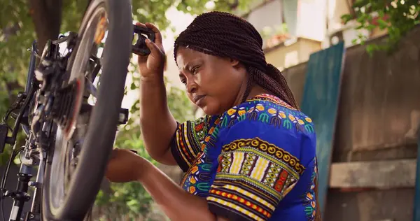 stock image Sportloving african american female checking and securing bicycle wheel with specialized multitool in home yard. Active black woman using expert work tool to tighten bike cogset for safe cycling.