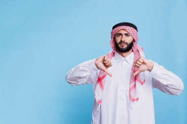 Geleneksel kıyafetli mutsuz müslüman adam iki baş parmak aşağı anlaşmazlık jesti portresi gösteriyor. Hoşnutsuz Arap kişi parmaklarıyla anlaşmazlık işareti gösteriyor ve kameraya bakıyor.