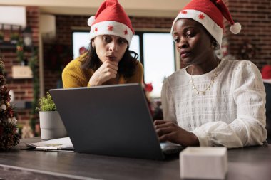 Noel tatilinde projenin son teslim tarihi için Noel Baba şapkası giyen meslektaşlar dizüstü bilgisayarda çalışıyor. Kurumsal şenlik döneminde süslü ofiste çalışan iki farklı kadın.