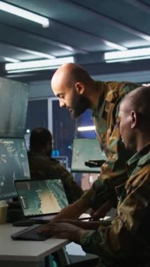 Görev Kontrol Merkezi 'ndeki dikey video askeri, meslektaşının yanındaki not defterini kullanarak ulusal güvenlik operasyonlarını denetliyor. Ordu uzmanlarından oluşan bir ekip gözlem odasındaki verileri inceliyor.