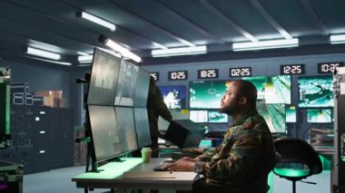 Askeri ajan kontrol odasında çalışıyor, hava keşif verilerini analiz ediyor ve meslektaşımla konuşuyor. İstihbarat ajanı ekranları kontrol ediyor, iş arkadaşı, kamera B ile tartışıyor.