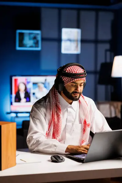 stock image Middle Eastern man in traditional attire working indoors, using a laptop for browsing while engaged in video call, using wireless headphones. Image shows male Arab person utilizing modern technology.