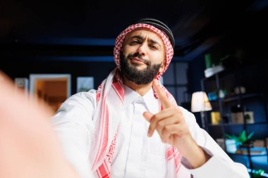 POV of a jovial Muslim man participating in a video conference. Middle Eastern guy wearing traditional clothing is vlogging online while holding a smart gadget and pointing at the camera. clipart