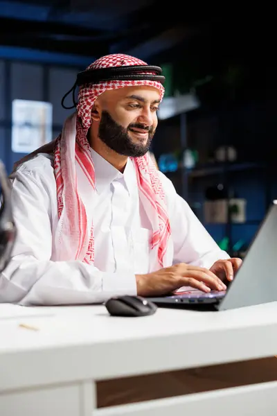 stock image While seated at his home office desk, a Middle Eastern male entrepreneur works effectively on his laptop. Portrait of Arab man checking e-mail, perusing the internet, and thoroughly examining notes.