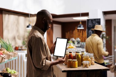 Afro-Amerikalı bir adam elinde kromakey şablonuyla tablet tutarken raflardaki ekolojik ürünleri, kavanozları ve bakkalları kontrol ediyor. Model için boş ekranlar. Sürdürülebilir yerel süpermarket.