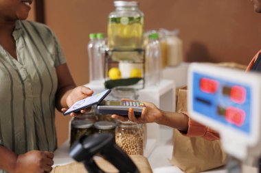 Afrika kökenli Amerikalı müşterinin, yerel marketteki taze organik ürünlere ödeme yapmak için cep telefonu cihazını pos makinesinde kullanması. Zenci kadın, biyolojik gıda ürünleri için cep telefonuyla NFC ödemesi yapıyor..