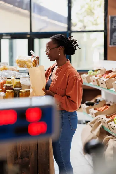 stock image Fusilli pasta is poured into a brown paper bag by black woman wearing glasses in grocery store. African American lady fills plastic free bag with freshly made spaghetti at bio food convenience shop.