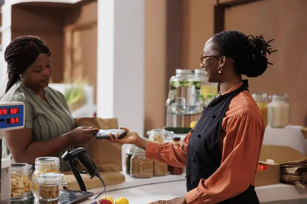 stock image African American woman happily makes cashless payment at market counter using credit card. She smiles as she communicates with the vendor and completes the transaction with a debit and pos machine.