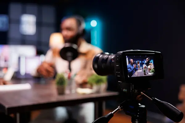 stock image Detailed view of professional camera filming a live talk show between two black men in home studio. Selective focus of recording equipment being used by african american male influencers.