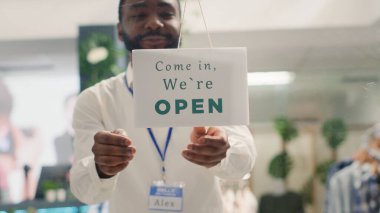 Smiling storekeeper finally reopening store after restocking elegant high end fashion boutique. Man turning closed sign from clothing store door after adding new collection, close up clipart