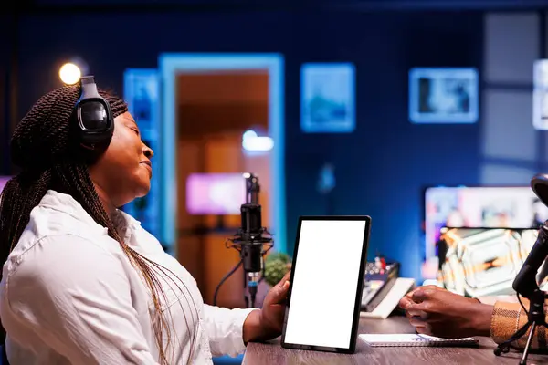 stock image Black woman grasping device displaying blank white screen while hosting a talk show. Female content creator using headphones and podcast equipment while holding tablet with chromakey template.