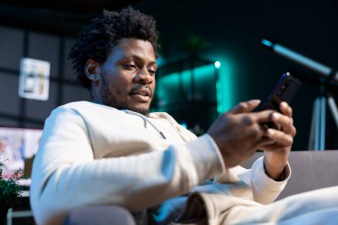 Neşeli adam mesajlaşarak rahatlıyor, buluşma saatini tartışıyor. BIPOC çalışanı modern şık oturma odasında rahat bir koltukta oturuyor cep telefonuyla yazı yazıyor, arkadaşıyla buluşacağı için heyecanlı.