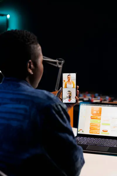 stock image Over the shoulder shot of freelancer using mobile device to communicate with others. African american woman using her cellphone to make a video conference call while sitting at a table with laptop.