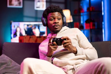 Afrika kökenli Amerikalı adam, rahat bir dairede video oyunu oynarken takım arkadaşlarıyla konuşmak için kulaklık kullanıyor. Oyuncu oyun konsolunda oyun oynarken çevrimiçi arkadaşlarıyla sohbet ediyor