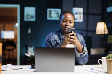 Afrika kökenli Amerikalı kadın blogcu masadaki dizüstü bilgisayarın önünde otururken cep telefonuyla mesaj atıyor. Genç siyahi kadın akıllı telefonunu tutuyor ve kullanıyor, internette ve sosyal medyada geziniyor..