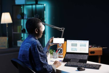 Beyaz ekranlı akıllı telefonu tutan ve dizüstü bilgisayar kullanan serbest çalışan bir kadın. Siyah kadın kişisel bilgisayarının önünde otururken yatay olarak cep telefonunu boş kromakey şablonuyla kavrıyor..