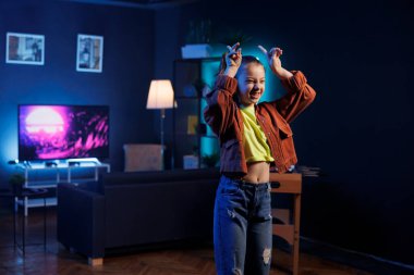 Çocuk oturma odasında üç boyutlu animasyonlar gösteren bilgisayar ekranlarıyla çevrili bir şekilde trend dans gösterisi yapıyor. Neon ışıklı ev stüdyosunda dans eden çocuk çevrimiçi hayranlar için içerik oluşturuyor.
