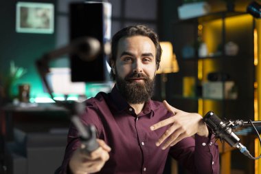 İçerik üreticisi cep telefonuyla konuşuyor. Ev stüdyosunun içindeki selfie çubuğuna bağlı, izleyicilerle sohbet ediyor. RGB aydınlatılmış dairede vlog kaydetmek için telefon kamerası kullanan bir adam