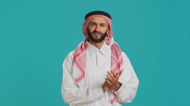 Geleneksel İslami kıyafetler giymiş Arap biri alkış işareti yapıyor, tebrik ediyor ve kendinden emin bir ifadeyle kameraya bakıyor. Müslüman adam stüdyoda destek veriyor ve alkışlıyor..