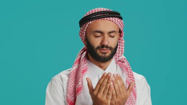 Müslüman biri kamerada Allah 'a dua ediyor, mavi arka planda dini ayinlere inanıyor. Orta Doğulu adam Tanrı 'ya meditasyonla, İslami kültürel kimlikle tapıyor..